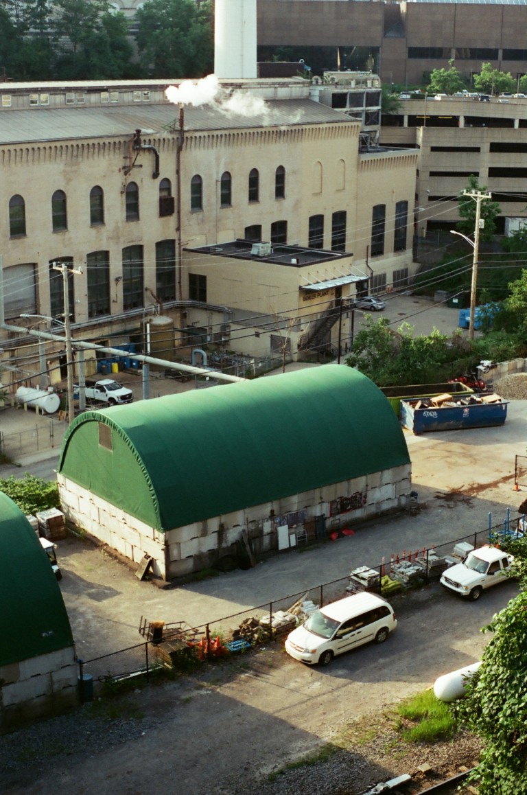 A view of an industrial area, with a tan building behind a green domed structure.
                        Two white cars can be seen at the front.