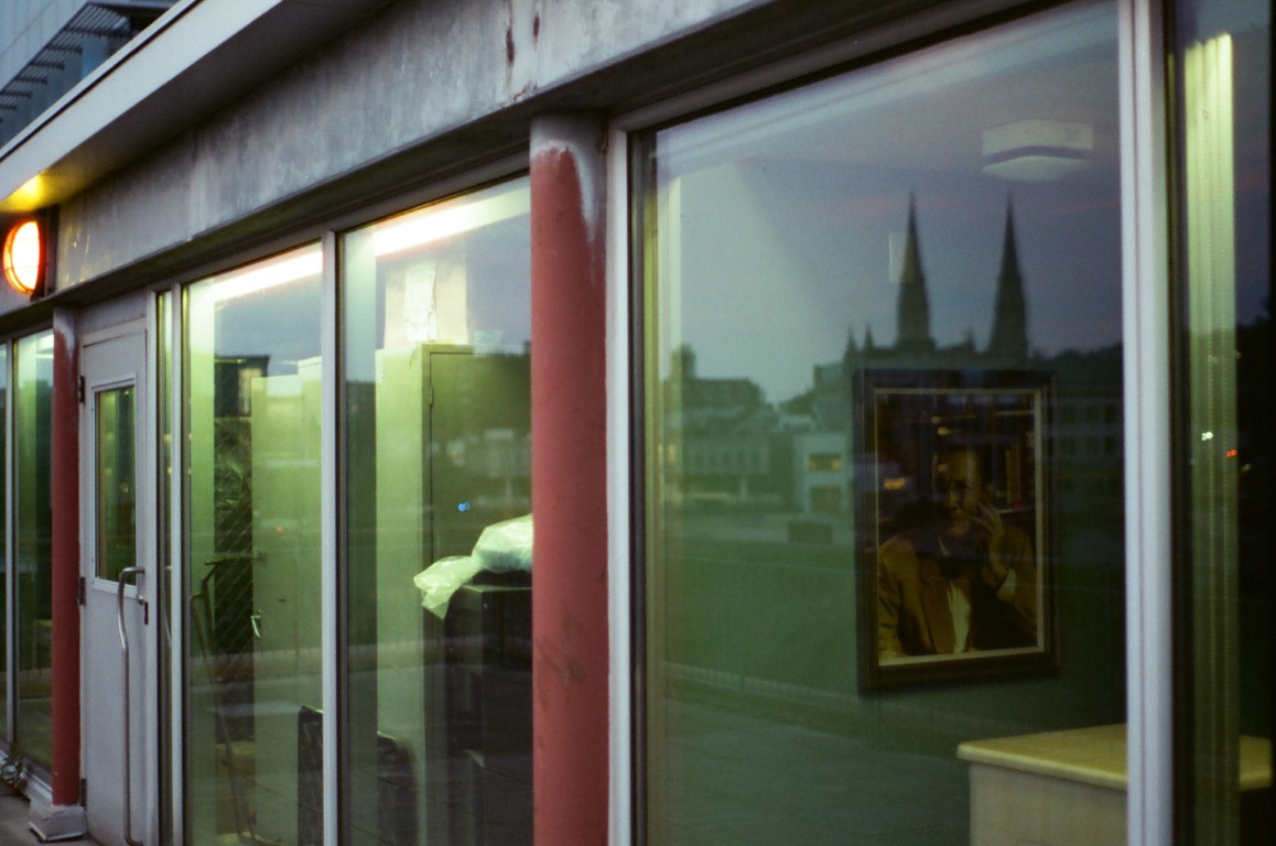 A set of windows outside of a building. The windows are reflecting a number of buildings.
                Inside the building, industrial shelves and a painting of a man can be seen.