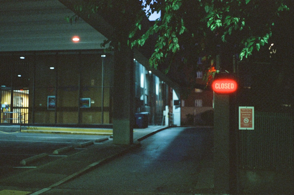 A nighttime scene of a building, in the foreground is a sign reading 'CLOSED'
                illuminated in red.