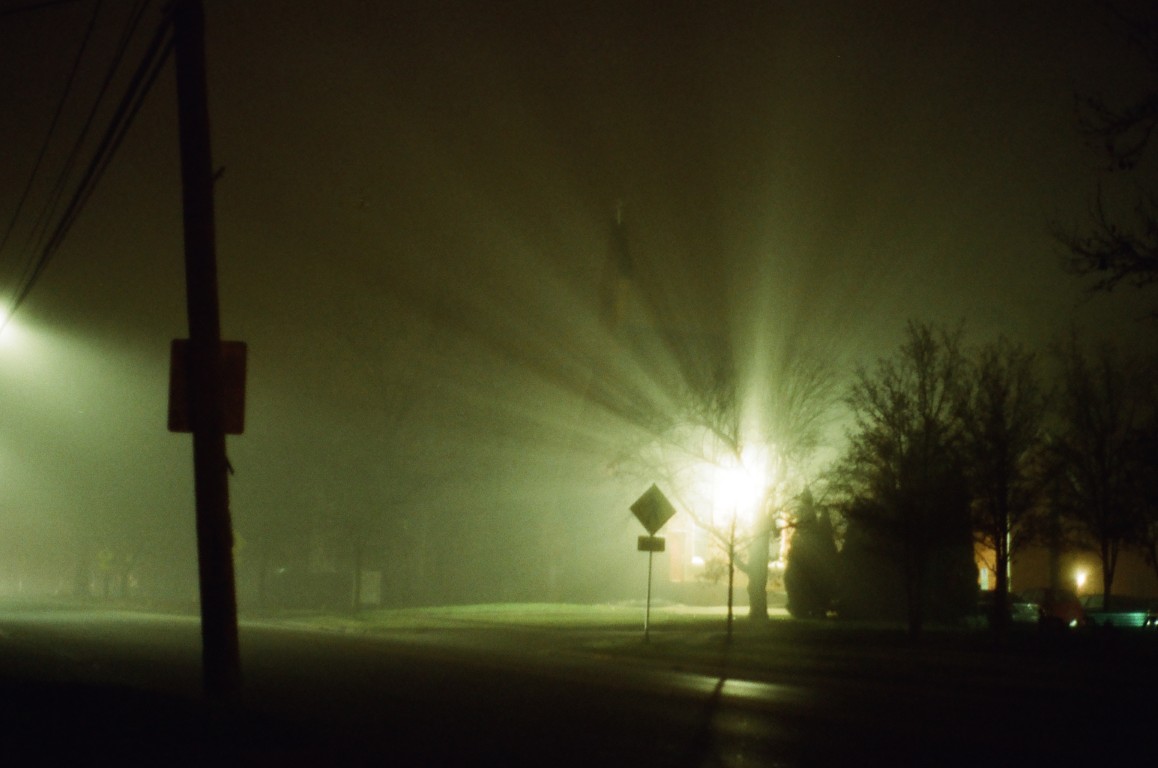Beams of light spill across a night scene, emanating from a church. Trees and a couple
                of road signs are outlined by the bright light.
