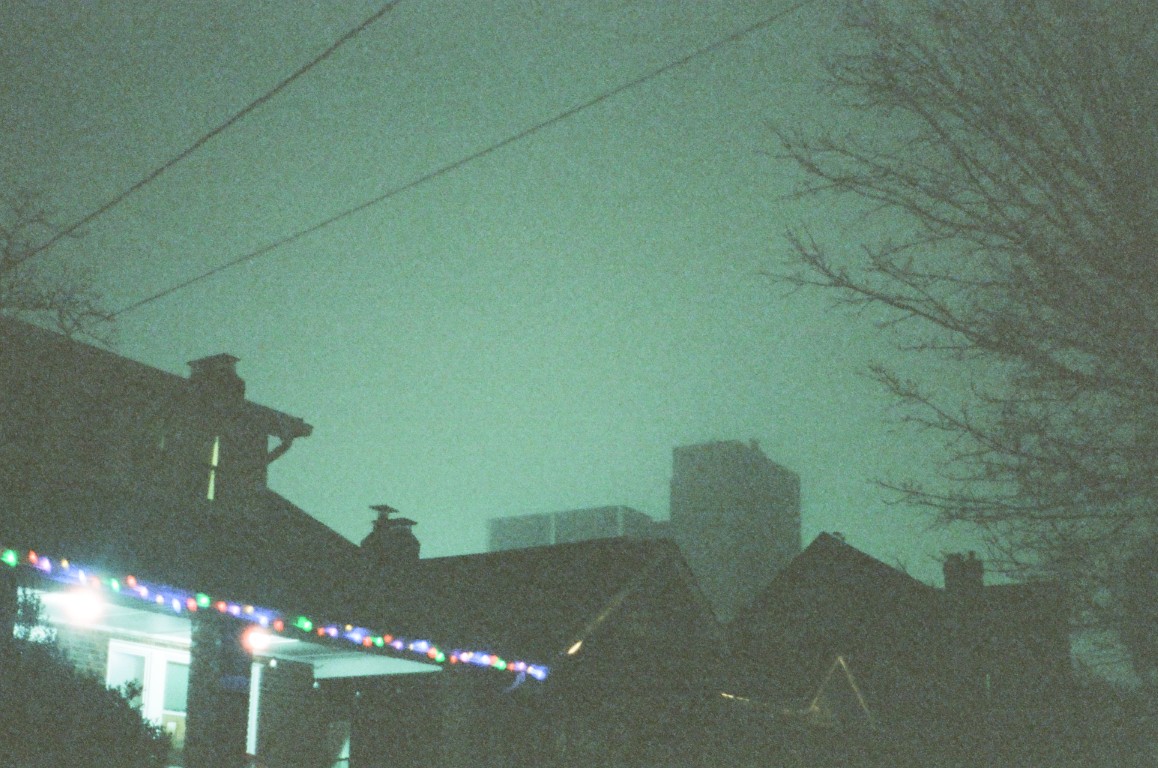 An eerie green light illuminates a foggy night, where the tops of houses and buildings can
                be seen. A row of Christmas lights runs along the edge of one roof.