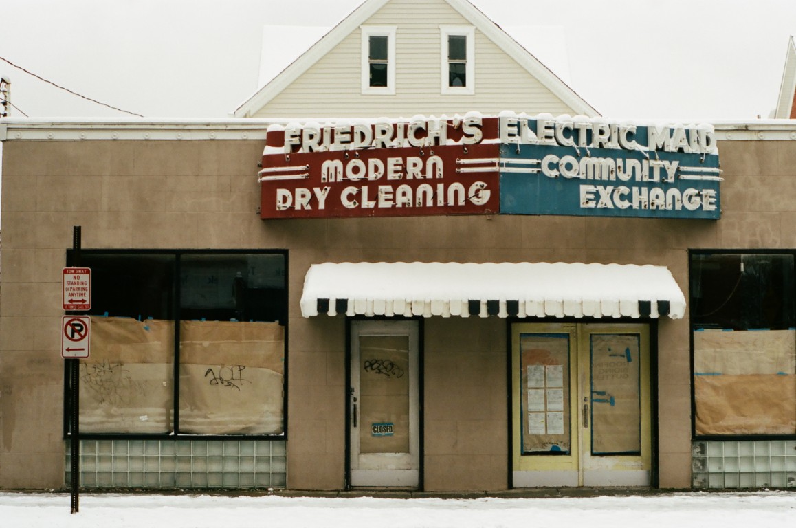 The front of a shuttered storefront. A large sign across the front is split into two halves.
                One half, in red, reads 'FRIEDRICH'S MODERN DRY CLEANING,' while the other half, in blue, reads
                'ELECTRIC MAID COMMUNITY EXCHANGE.'