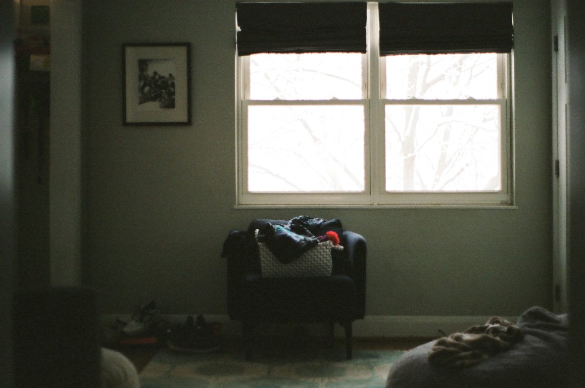 A scene of a living room, with various objects scattered around. Bright white light comes
                from a pair of windows, and a chair in the center of the scene has a basket sitting on it, with
                a red hat poking out.