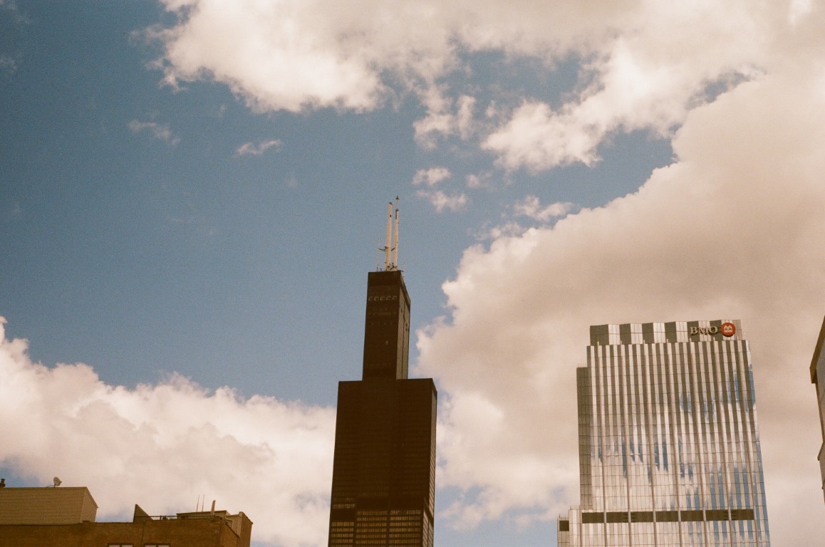 Three buildings against a blue sky with white clouds. The first building, barely visible
                on the left, is a square, brown, brick building. The second building is the Willis Tower in
                Chicago, a black, blocky skyscraper with white antennas. The final building, on the right,
                is a shiny glass building with wavy sides and a sign that reads 'BMO.'
